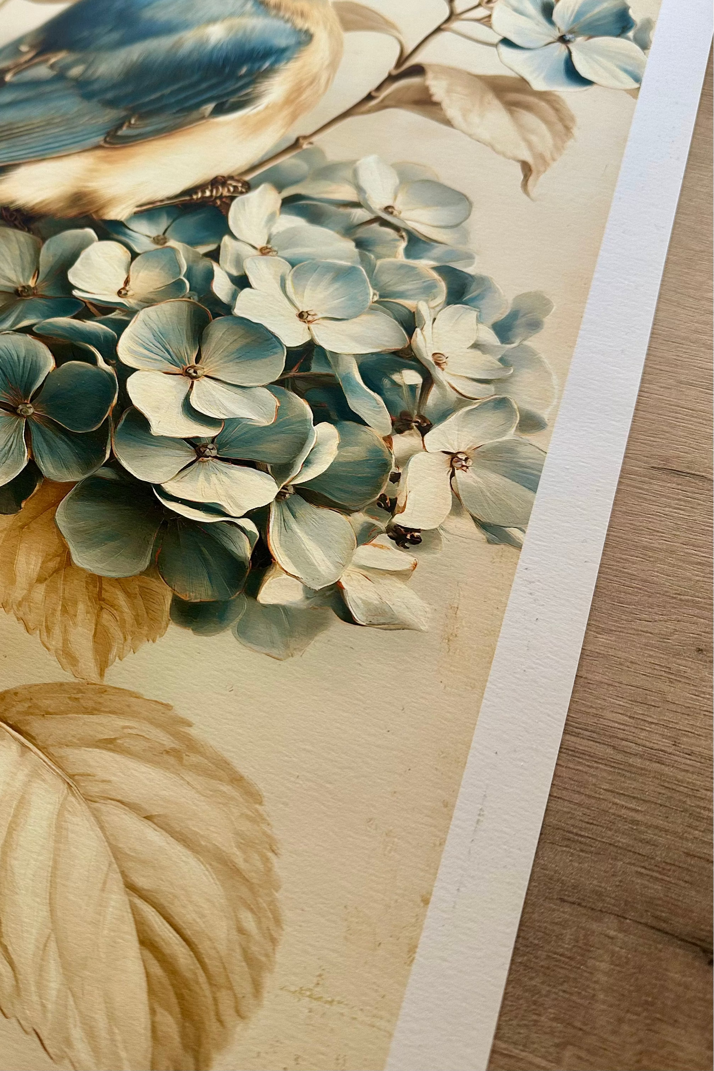 A close-up of a printed artwork showing a bluebird perched on a branch with pale blue hydrangea flowers and beige leaves, photographed on a light wood surface.