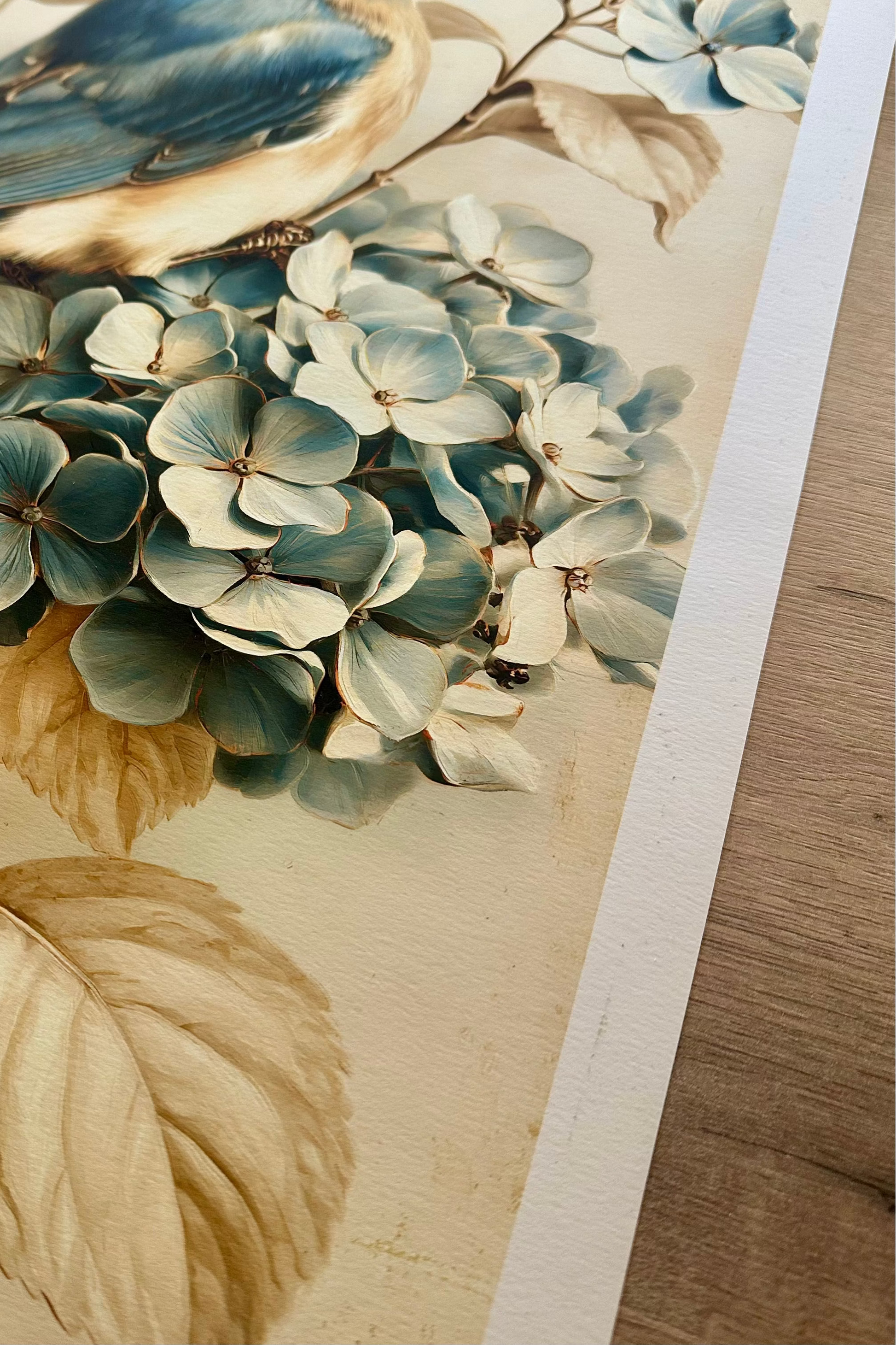 A close-up of a printed artwork showing a bluebird perched on a branch with pale blue hydrangea flowers and beige leaves, photographed on a light wood surface.